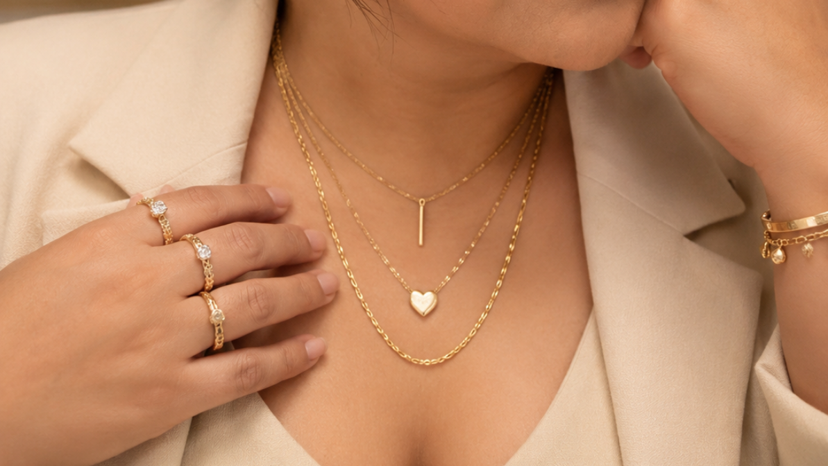 A woman with dark hair in a bun wearing layered gold necklaces, hoop earrings, and rings, posing in a cream-colored blazer against a warm, softly lit background.